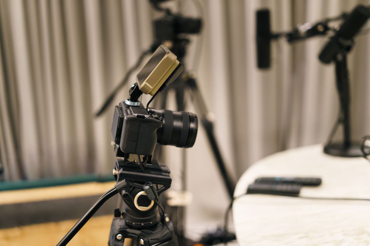 Cámara de vídeo profesional montada en un trípode dentro de un pequeño estudio de grabación, con micrófonos de brazo al fondo